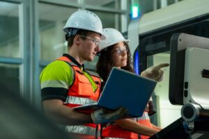 Two workers, wearing white hard hats, safety glasses, and high-visibility vests, stand in a factory or industrial setting.